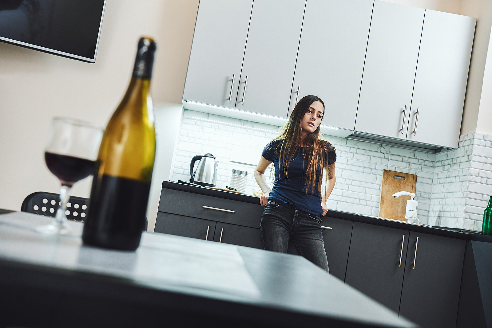 A young woman stands in her kitchen, and open bottle of wine and a full wine glass on the table. She looks distressed. Relapse, rehab, alcohol, addiction.