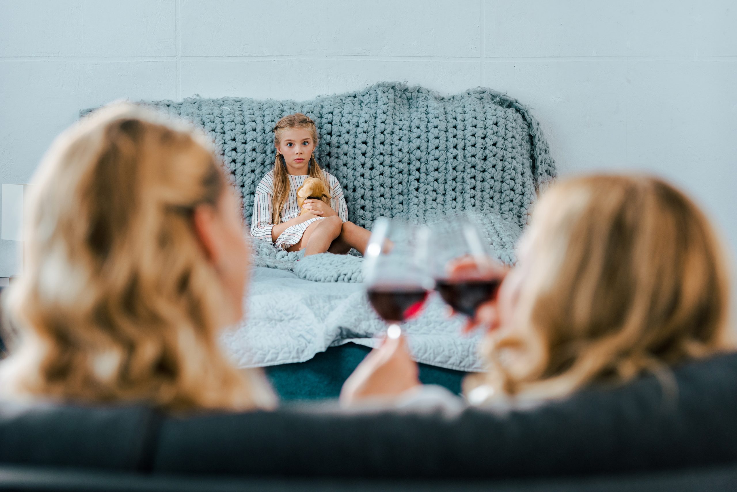 sad lonely child sitting on bed with toy while her mother clinking glasses of wine with friend on foreground